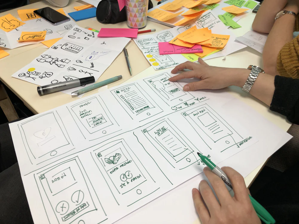 Mains travaillant sur des croquis et maquettes papier autour d’une table de création